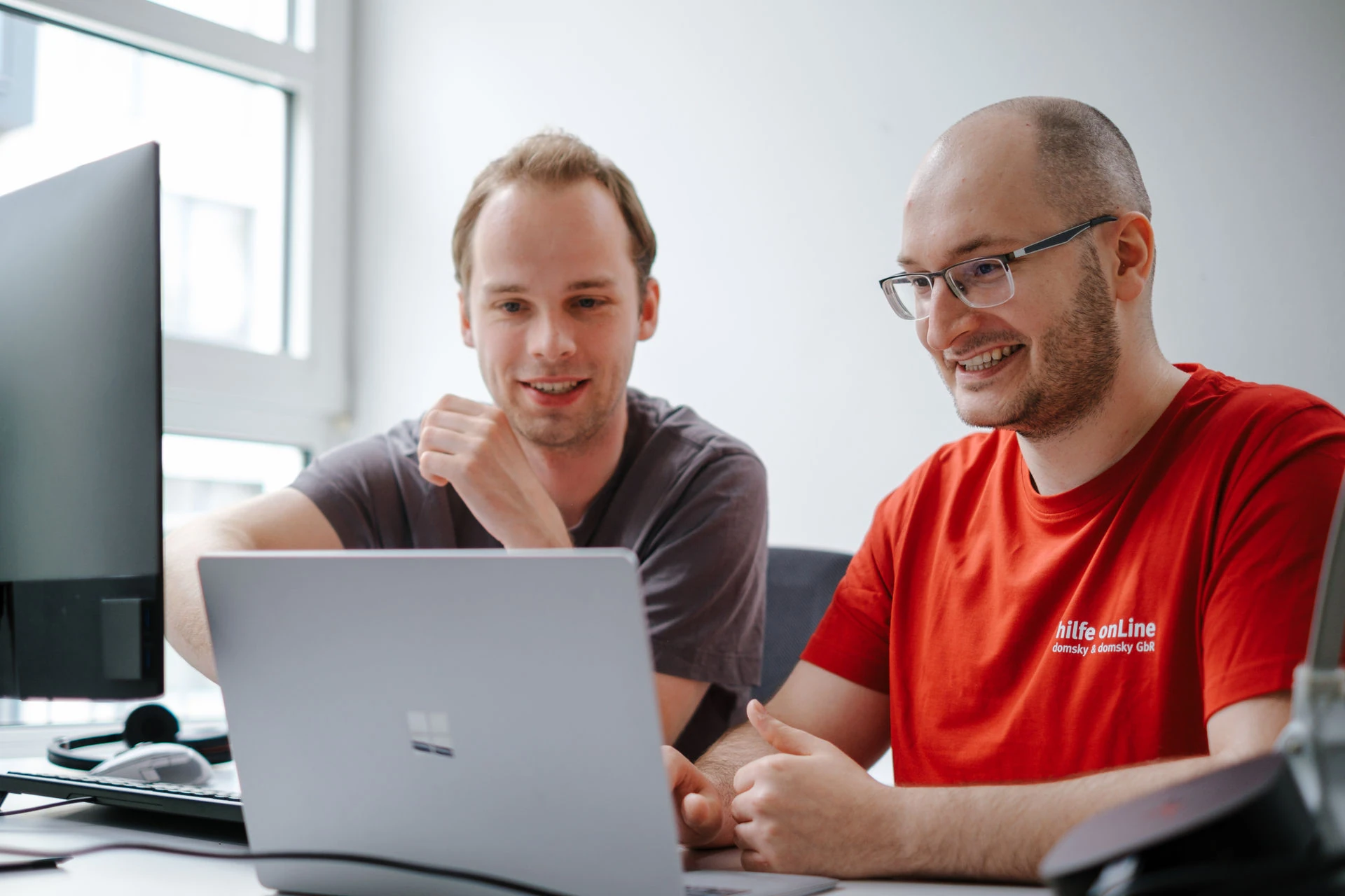 Zwei Männer sitzen an einem Schreibtisch und schauen gemeinsam auf den Bildschirm eines Laptops; einer zeigt auf den Bildschirm, beide wirken konzentriert und vertieft in ein Gespräch.