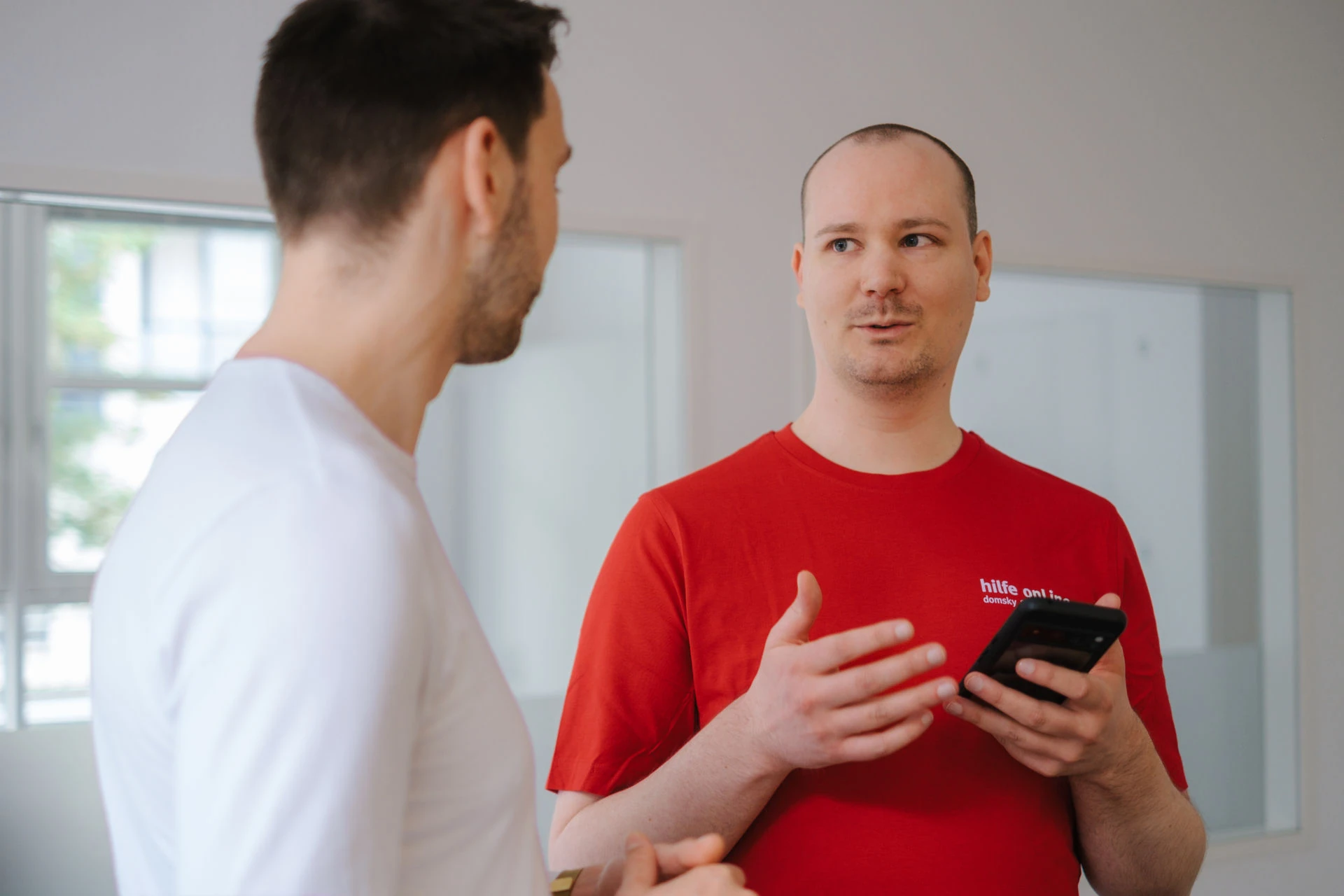 Zwei Männer stehen in einem Raum und unterhalten sich; der eine, der ein weißes T-Shirt trägt, hört zu, während der andere, der ein rotes T-Shirt trägt, mit der Hand gestikuliert und ein Smartphone in der Hand hält.