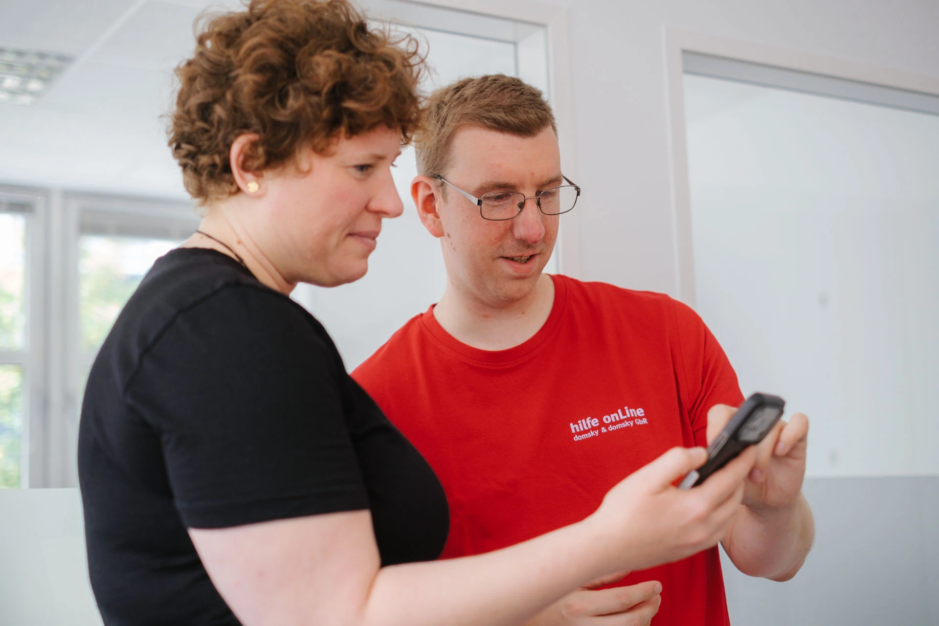 Zwei Personen stehen in einem Raum und schauen sich gemeinsam ein Smartphone an; die eine trägt ein schwarzes T-Shirt, die andere ein rotes T-Shirt mit der Aufschrift „hilfe onLine.“.