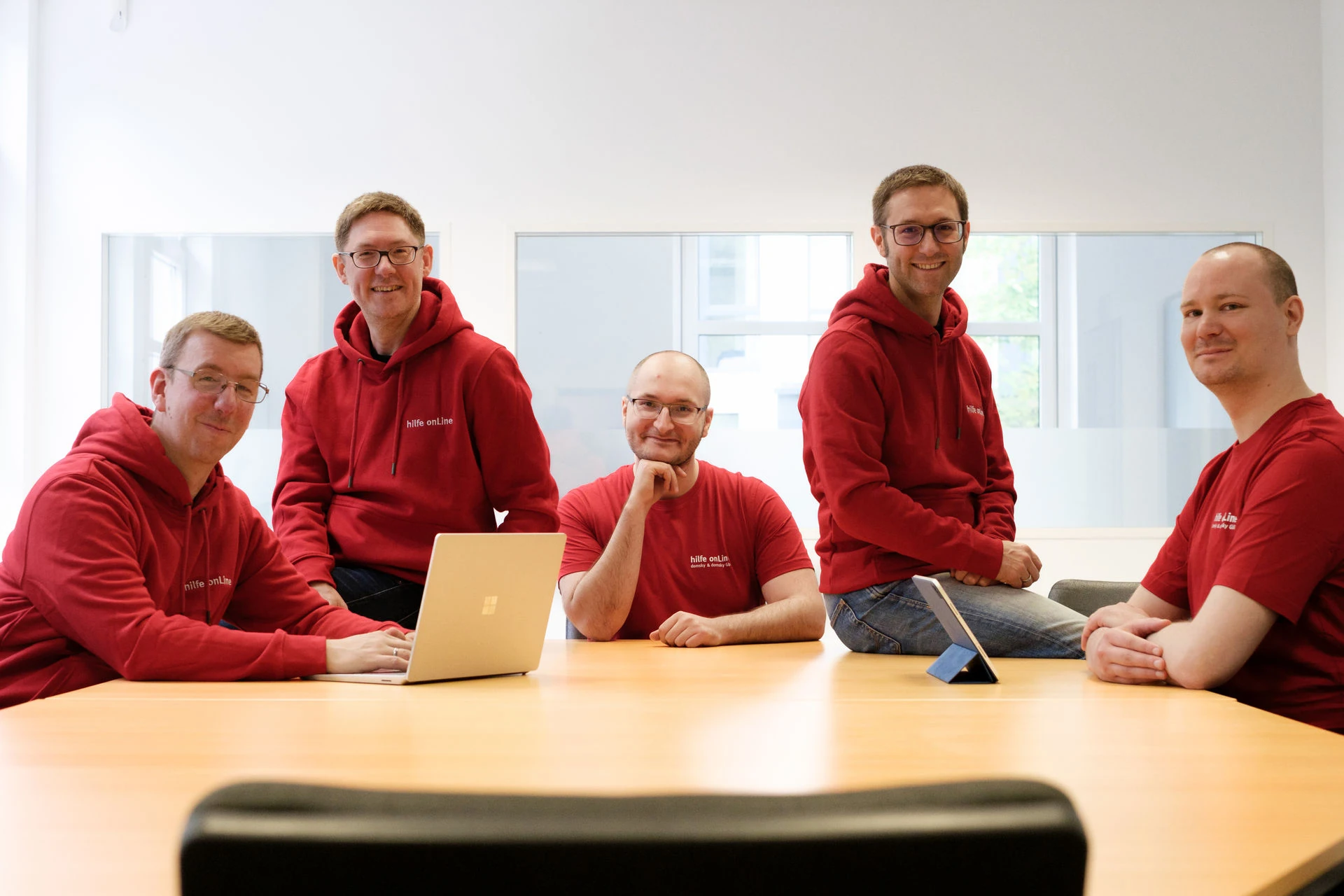 hilfe-online-teamfoto-it-unternehmen-bremen Fünf Männer in einheitlichen roten Kapuzenpullis sitzen und stehen in einem hellen Büroraum um einen Konferenztisch herum, auf dem ein Laptop und ein Tablet liegen.