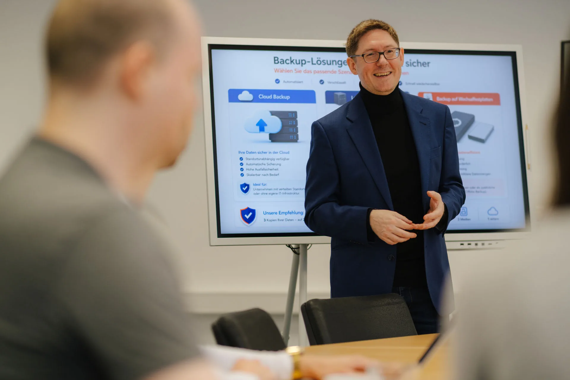 Ein Mann in einem dunklen Jackett präsentiert zwei Personen, die an einem Konferenztisch sitzen, auf einem Bildschirm Informationen zu Backup-Lösungen.