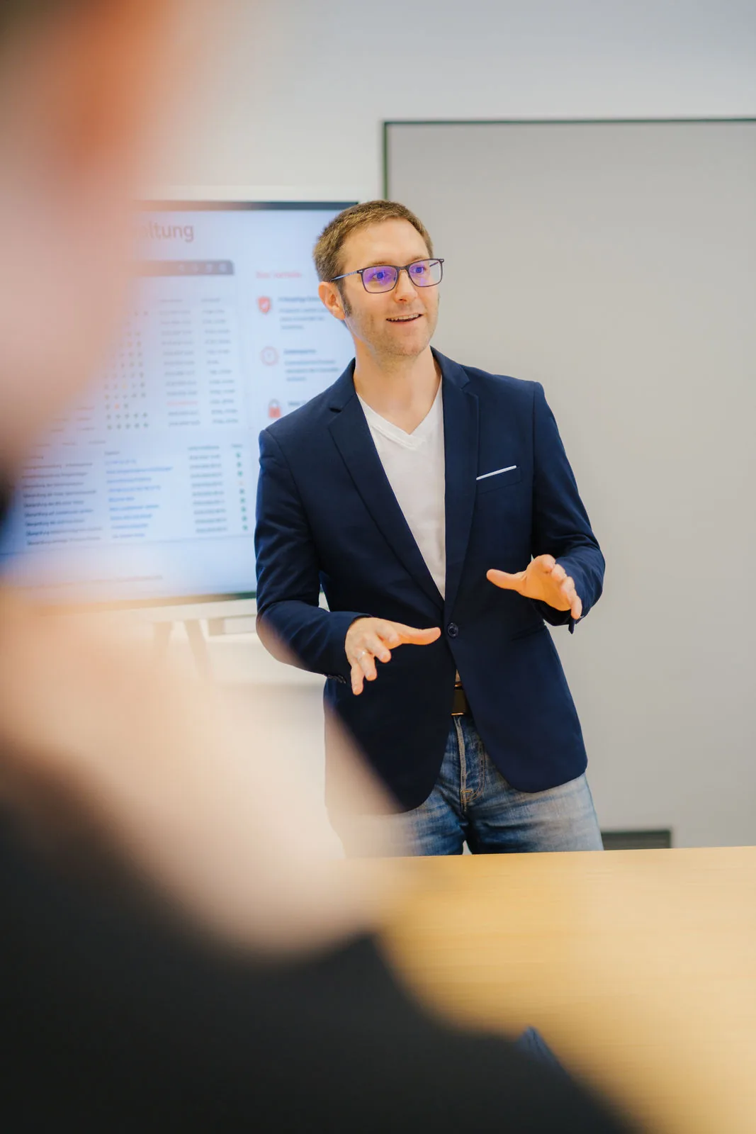Ein Mann in einem marineblauen Blazer und mit Brille steht neben einem Tisch, gestikuliert beim Sprechen, im Hintergrund ist eine Präsentationsleinwand zu sehen.
