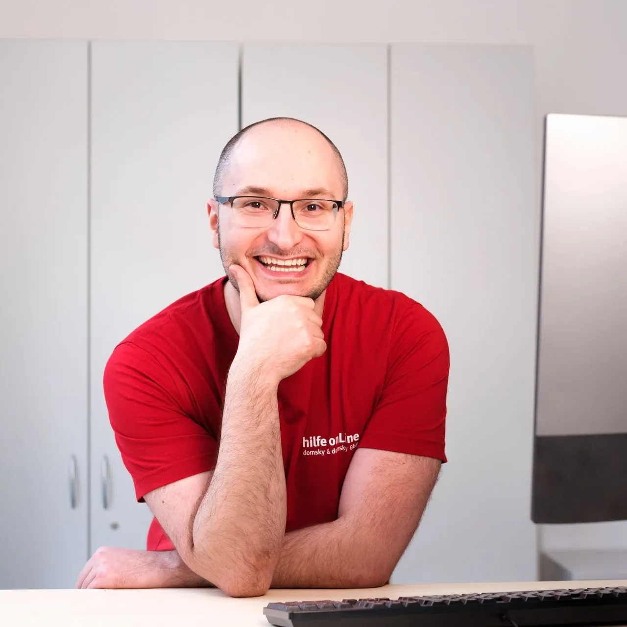 hilfe-online_bremen_Kevin-Rischka_Fachinformatiker-Systemintegration Eine Person mit Brille und rotem T-Shirt sitzt an einem Schreibtisch, lächelt und stützt ihr Kinn auf eine Hand. Im Hintergrund sind ein Computermonitor und weiße Schränke zu sehen.
