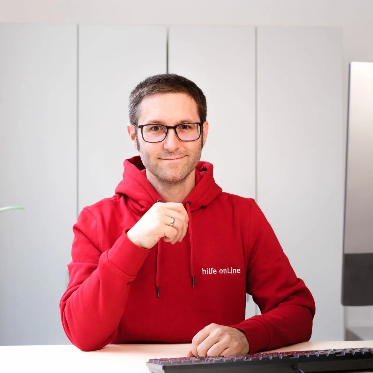 hilfe-online_bremen_Malte-Domsky_Geschaeftsleitung Eine Person in einem roten Kapuzenpulli und mit Brille sitzt an einem Schreibtisch vor einer Tastatur und lächelt in die Kamera.