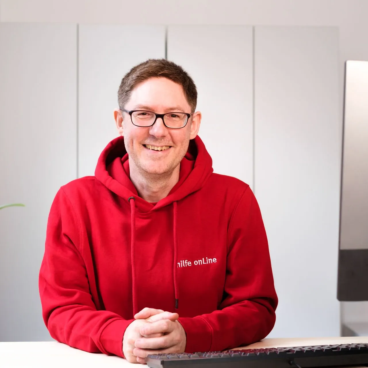 hilfe-online_bremen_Oliver-Domsky_Geschaeftsleitung Eine Person mit Brille und rotem Kapuzenpullover sitzt an einem Schreibtisch mit einer Tastatur und lächelt in die Kamera. Im Hintergrund sind ein Computermonitor und eine Pflanze zu sehen.