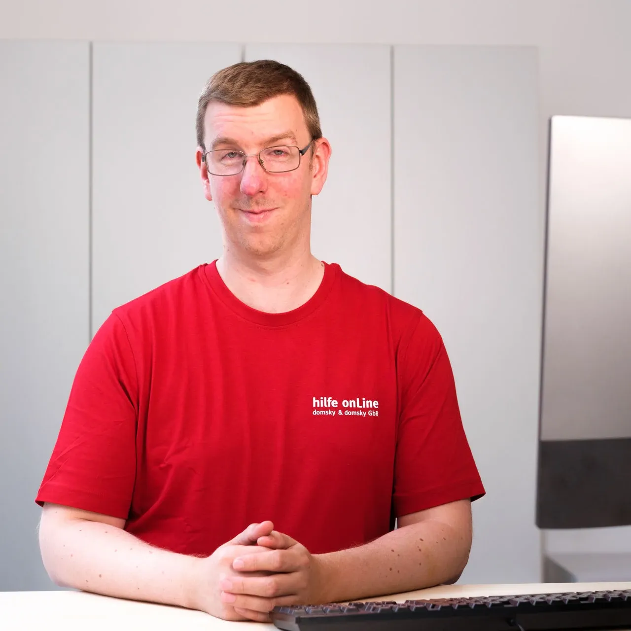hilfe-online_bremen_thiemo-eickel_Fachinformatiker-Systemintegration Ein Mann in einem roten T-Shirt mit der Aufschrift „hilfe online“ sitzt mit verschränkten Händen an einem Schreibtisch, vor sich eine Computertastatur.
